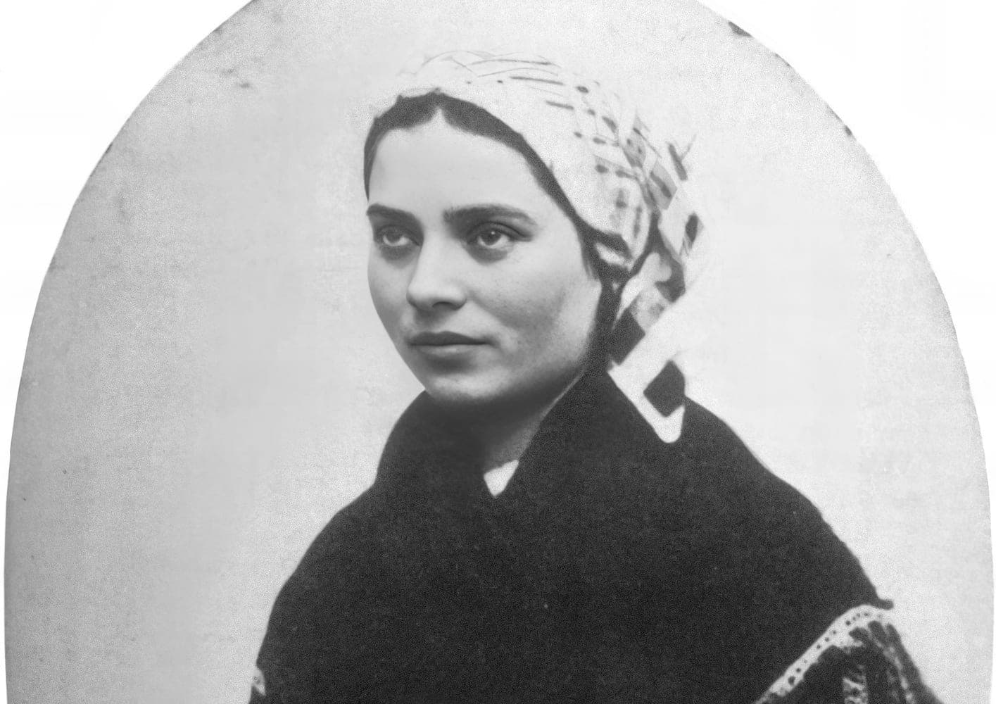 A photograph of Bernadette Soubirous taken in 1863, a young woman in a dark shawl and white headscarf, her face calm and direct