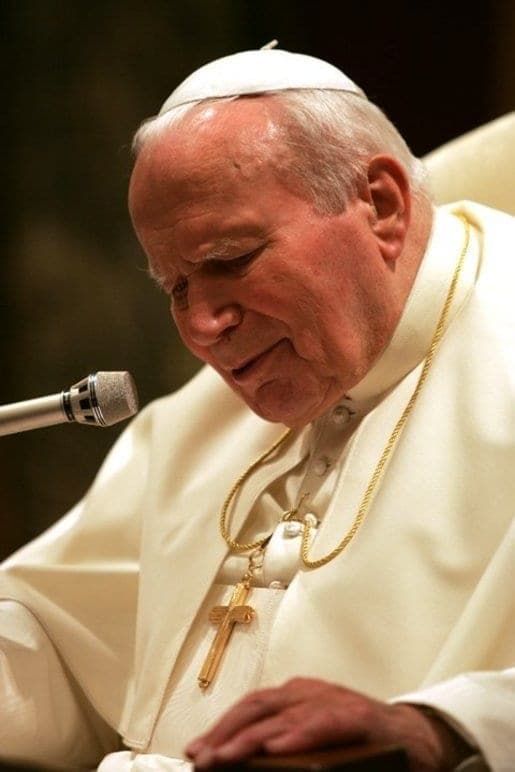 A photograph of Pope John Paul II in white papal dress and zucchetto, head bowed in prayer over a microphone, a golden pectoral cross at his breast