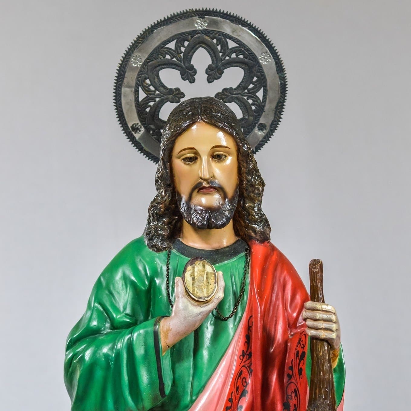 A polychrome statue of St. Jude Thaddeus in a green tunic and red mantle, holding up the oval medallion of Christ that hangs from his neck, a wooden club in his other hand and a pierced metalwork halo behind his head