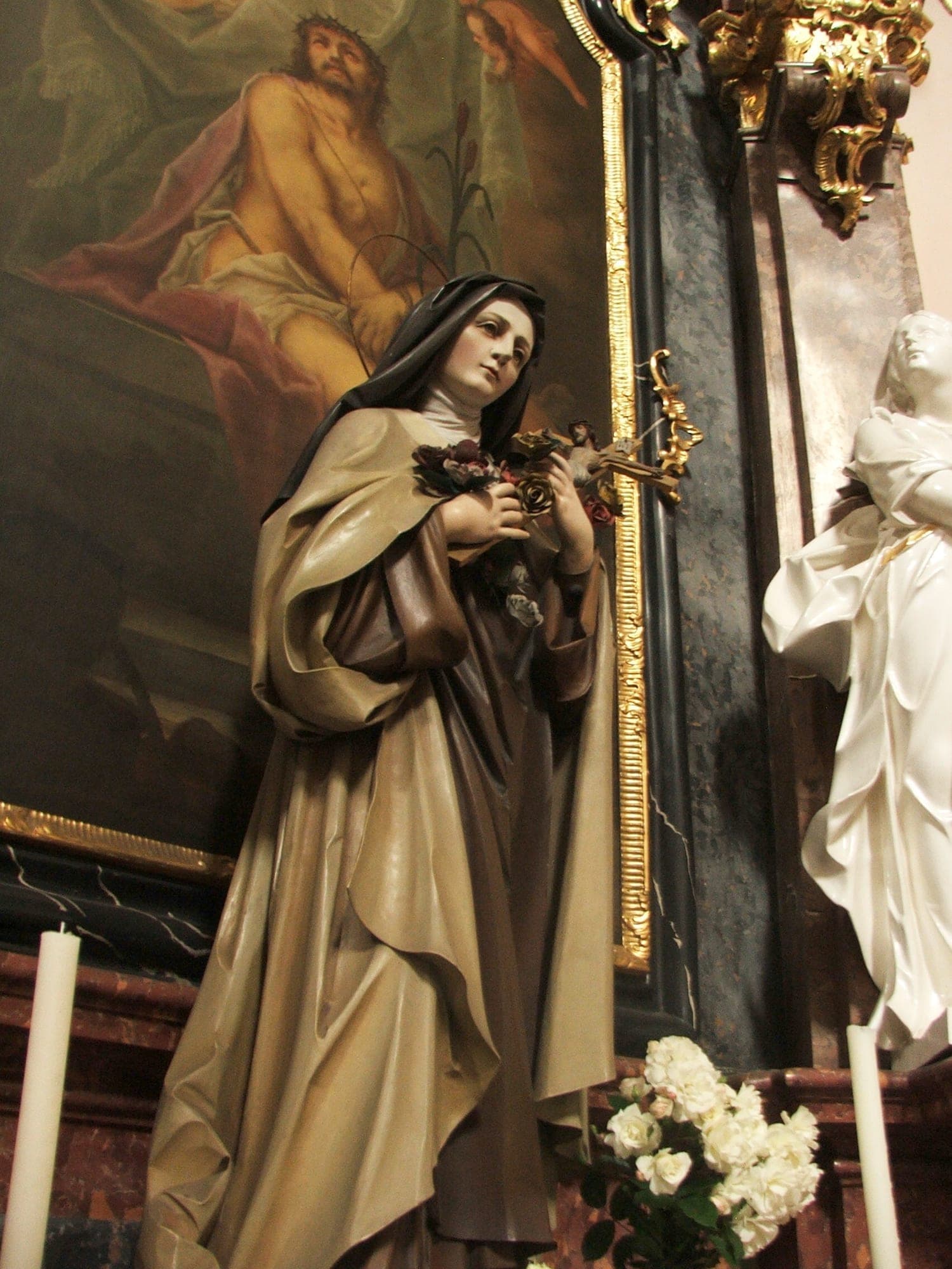 A statue of St. Thérèse with roses and crucifix standing in a chapel beneath a painting of Christ crowned with thorns